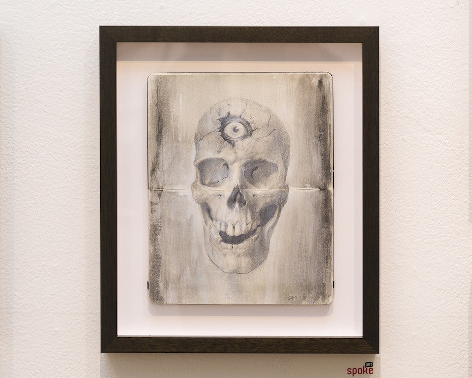 Cody Miles presents a framed artwork of a skull with a third eye, using graphite and acrylics on a textured backdrop. The black frame stands out against the white wall, evoking an eerie allure akin to Moleskine sketches.