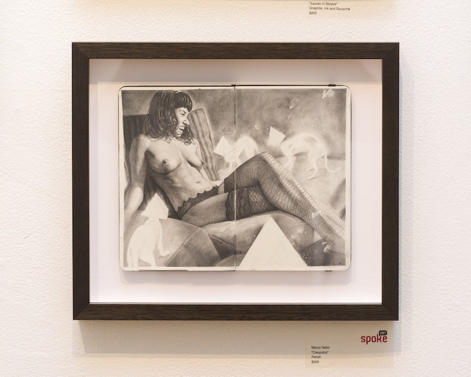 A framed Marco Nelor pencil drawing on Moleskine depicts a provocatively reclining figure reminiscent of Cleopatra in lingerie and stockings, mounted on a white wall with spoke labeled at the bottom right of the matting.