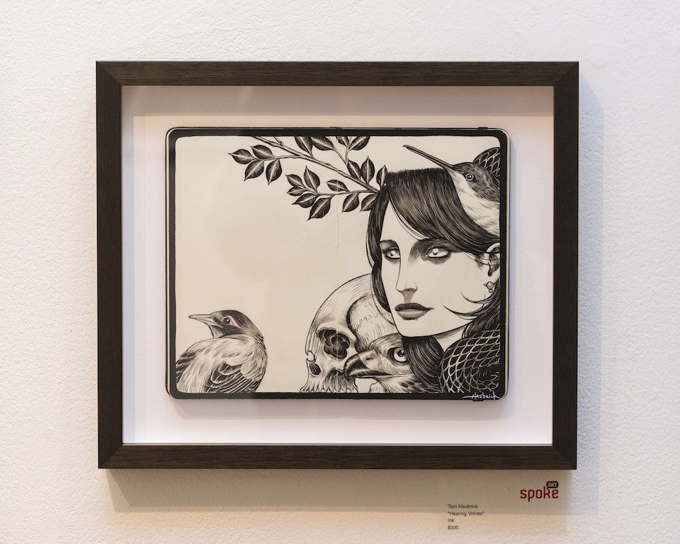 Framed artwork by Tom Haubrick features a woman, crows, a skull, and a leafy branch in ink on Moleskine. Elegantly presented in a dark wooden frame with a gallery label.