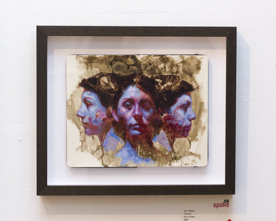 A framed artwork by John Wentz depicts a surreal portrait of a woman with multiple faces, expertly painted in oil on Moleskine. Brown, purple, and blue hues blend with the abstract background and central figure on a pristine white wall.