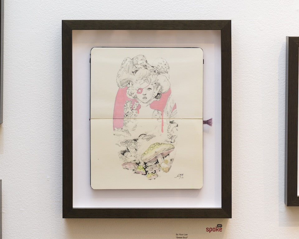 Framed mixed media art by So Youn Lee showcases a person with intricate hair and clothing in pastel hues against a pink backdrop, all set in a black frame on a white wall.
