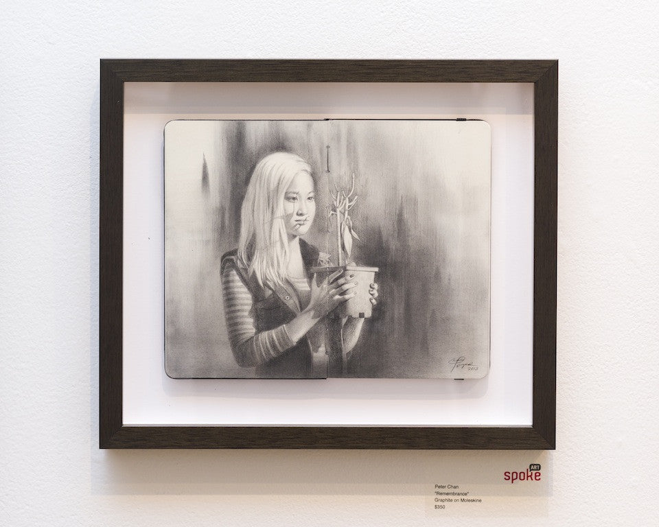 Peter Chans graphite artwork on Moleskine features a grayscale blend of realistic and sketchy styles, showing a woman with long hair holding a potted plant. It hangs on a white wall with a small remembrance label at the bottom right of the frame.