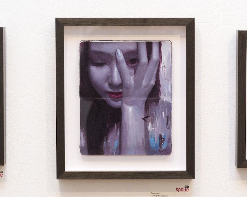 Peter Chans framed oil painting on Moleskine portrays a woman with her face partly hidden by her hand, emphasizing red lips and closed eyes amid a purple and red color scheme. This evocative artwork invites viewers to explore new perspectives in beauty when displayed on a gallery wall.