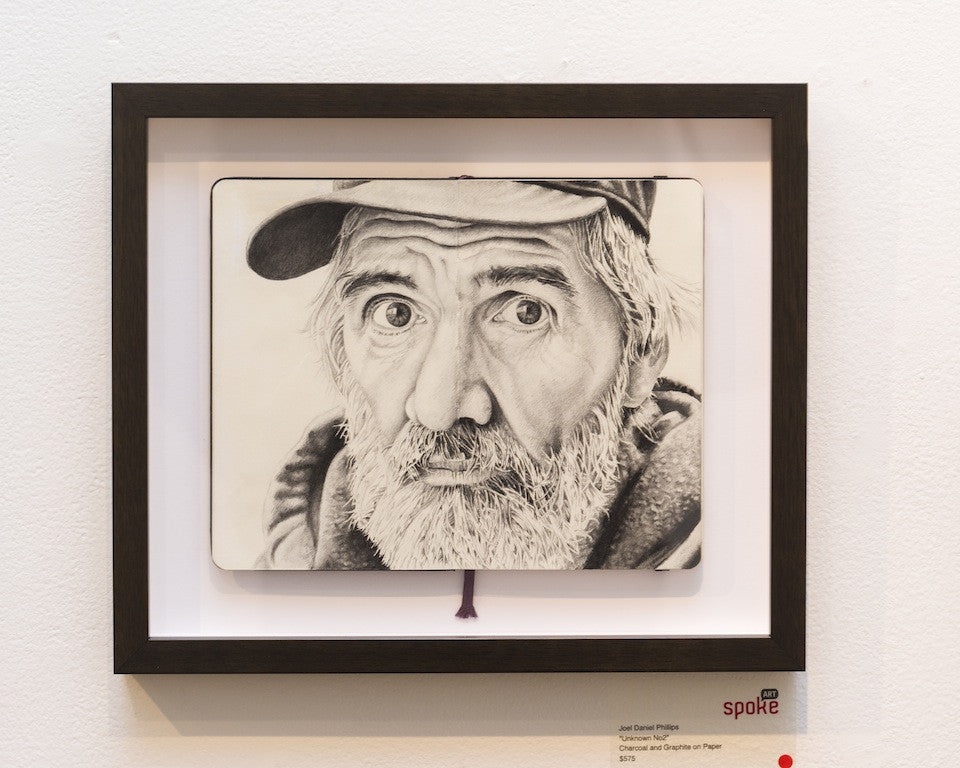 A framed pencil sketch titled Framed Charcoal and Graphite Portrait by Joel Phillips features an elderly man with a beard and cap, wide eyes, and a thoughtful expression. Displayed on a gallery wall, this exquisite piece is labeled as art for sale in the bottom corner.