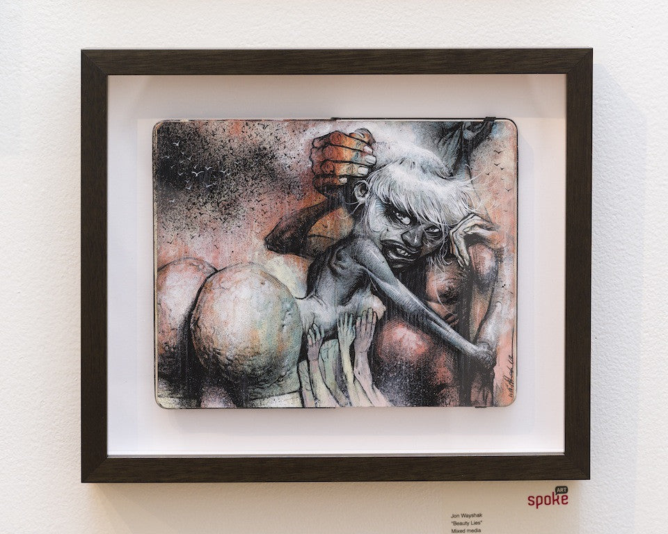 Framed artwork by Jon Wayshak showcasing a fantastical creature with human-like features and a mischievous expression, surrounded by abstract shapes and hands. This mixed media piece blends muted colors and textures against a white wall.