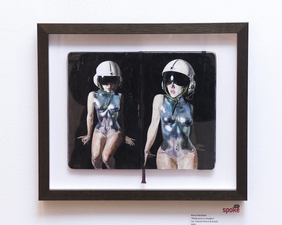 Kemp Remillards framed artwork features two figures in metallic bodysuits and helmets, artistically rendered with colored pencil on sketchbook pages and displayed on a sleek white wall.