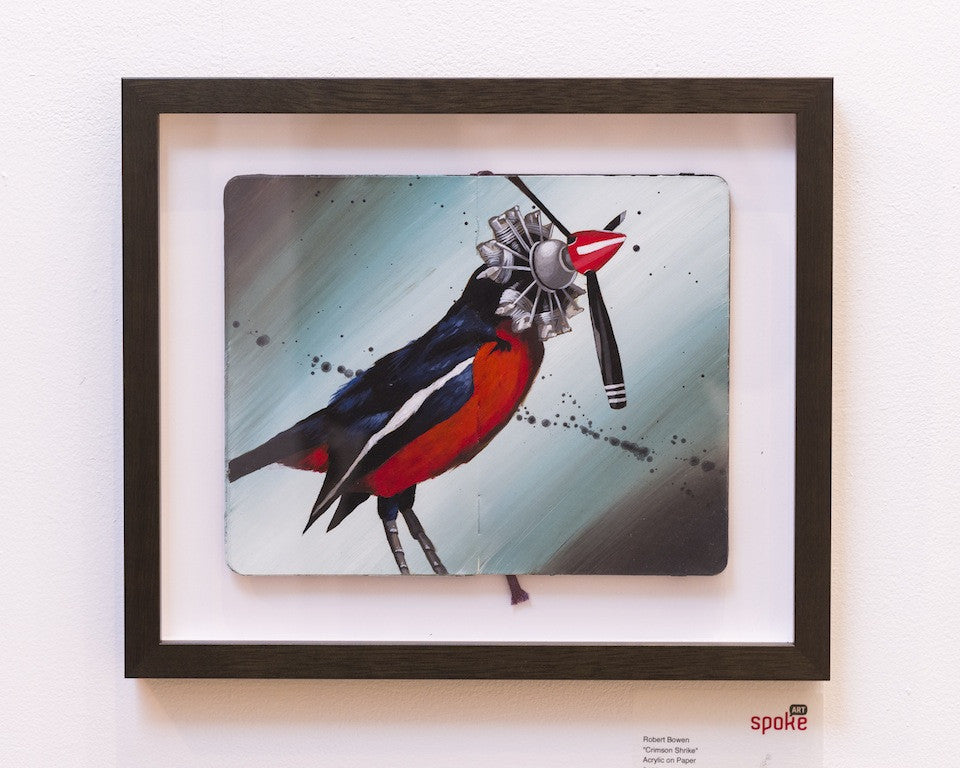 Framed artwork, Crimson Shrike by Robert Bowen, showcases a bird with a mechanical head and propeller. Painted in acrylic on paper, its vivid red and blue feathers stand out against a blurred backdrop, mixing surrealism and imagination by blending nature with technology.