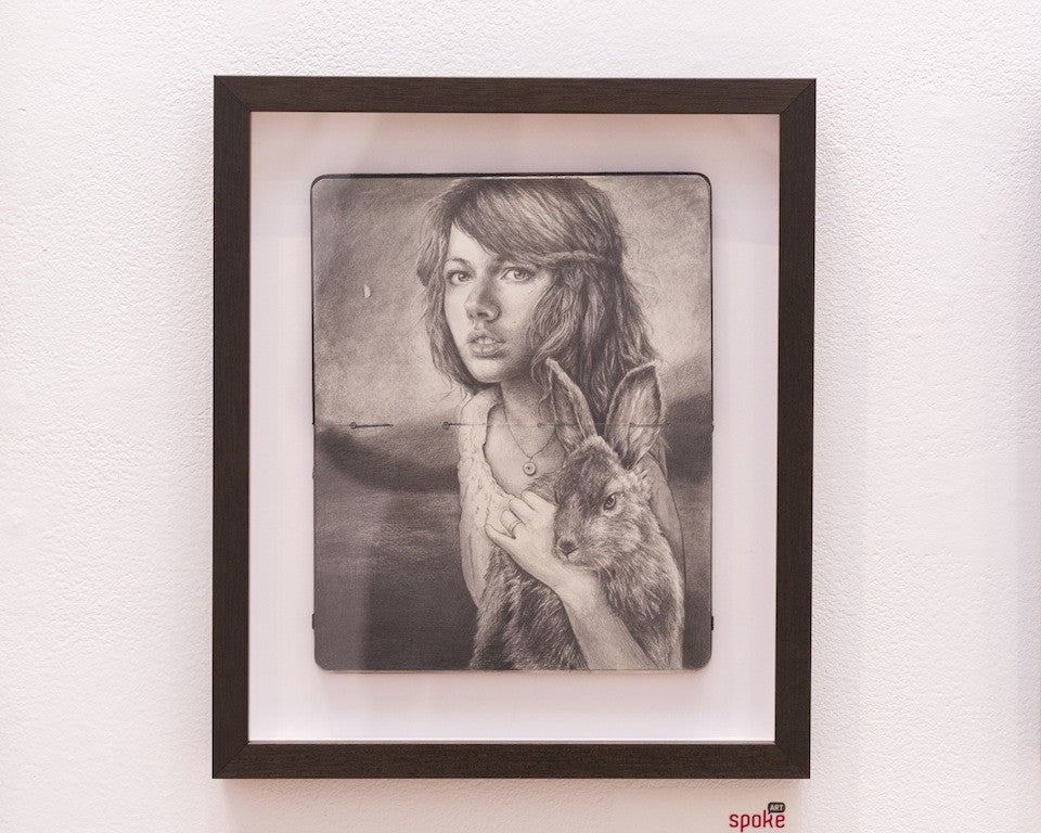 A Michael Shapcott artwork features a woman with medium-length hair holding a rabbit. Sketched in graphite on Moleskine, the detailed black-and-white piece captures a serene pose and is framed against a white wall.