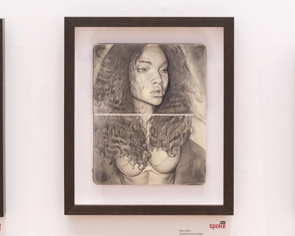 A Marco Nelor framed pencil portrait of a woman with long curly hair showcases detailed realism. Set against a white wall, this monochromatic artwork beautifully mirrors an Ashley study in its exquisite depiction of her face and upper body.