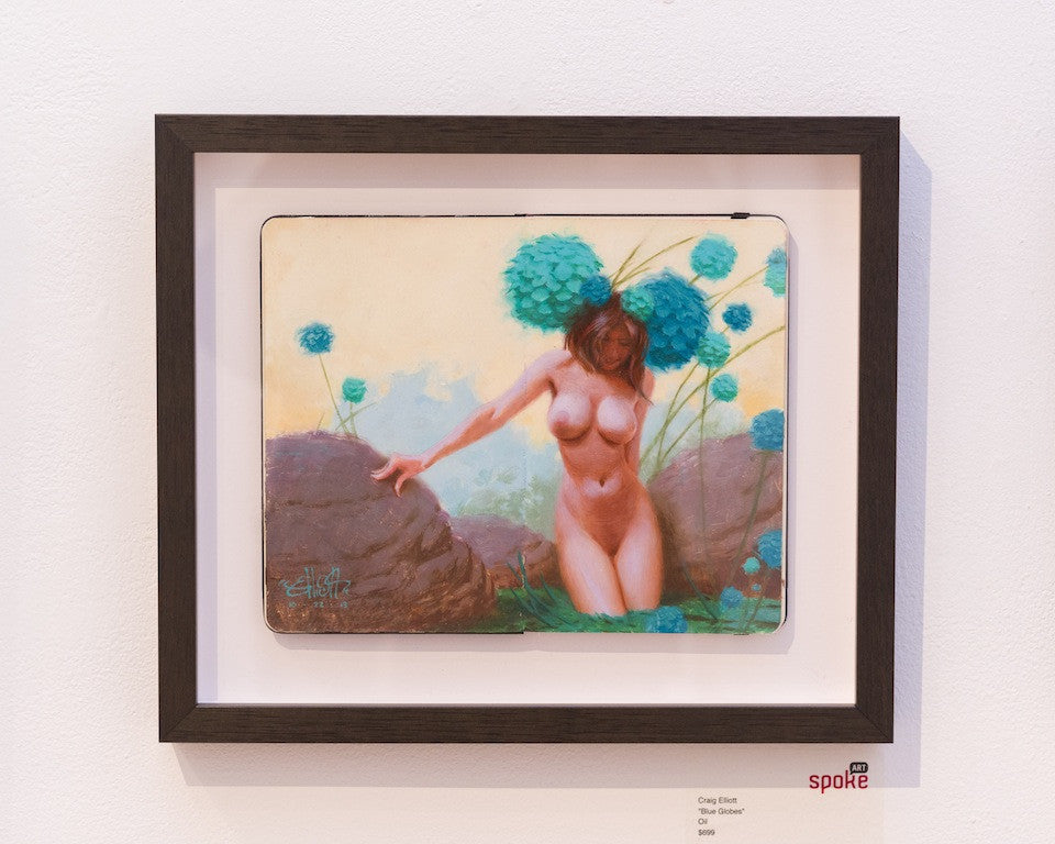 A framed oil painting by Craig Elliott depicts a nude woman with large turquoise flowers and rocks against a light background, beautifully showcased on a gallery wall.