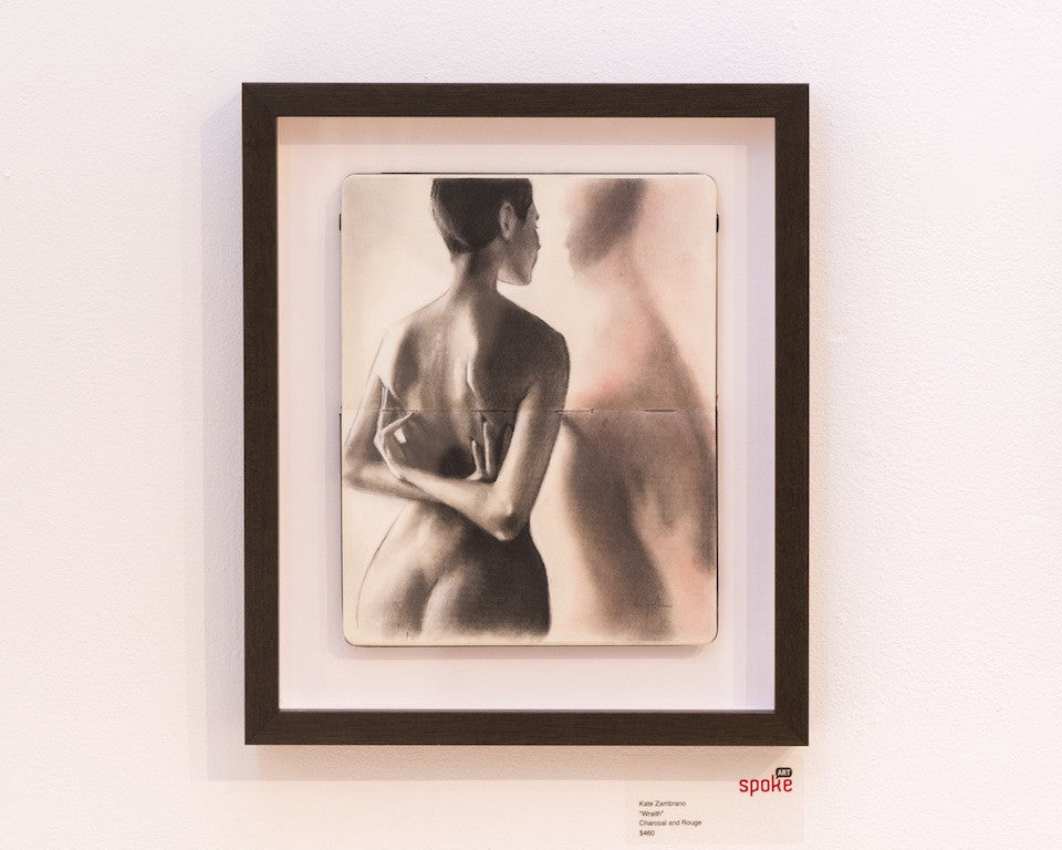 This framed charcoal drawing by Kate Zambrano features a nude figure with back turned and arms partially crossed, creating a wraith-like presence in the blurred mirror reflection. Displayed on a white wall with a label below, it captivates with its haunting elegance.
