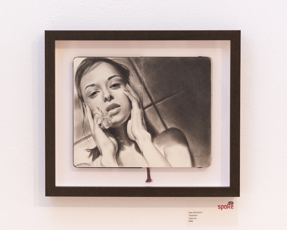 Kate Zambranos framed charcoal drawing on Moleskine depicts a woman with a thoughtful expression holding a flower, set in an obsidian frame against a white wall, labeled underneath.