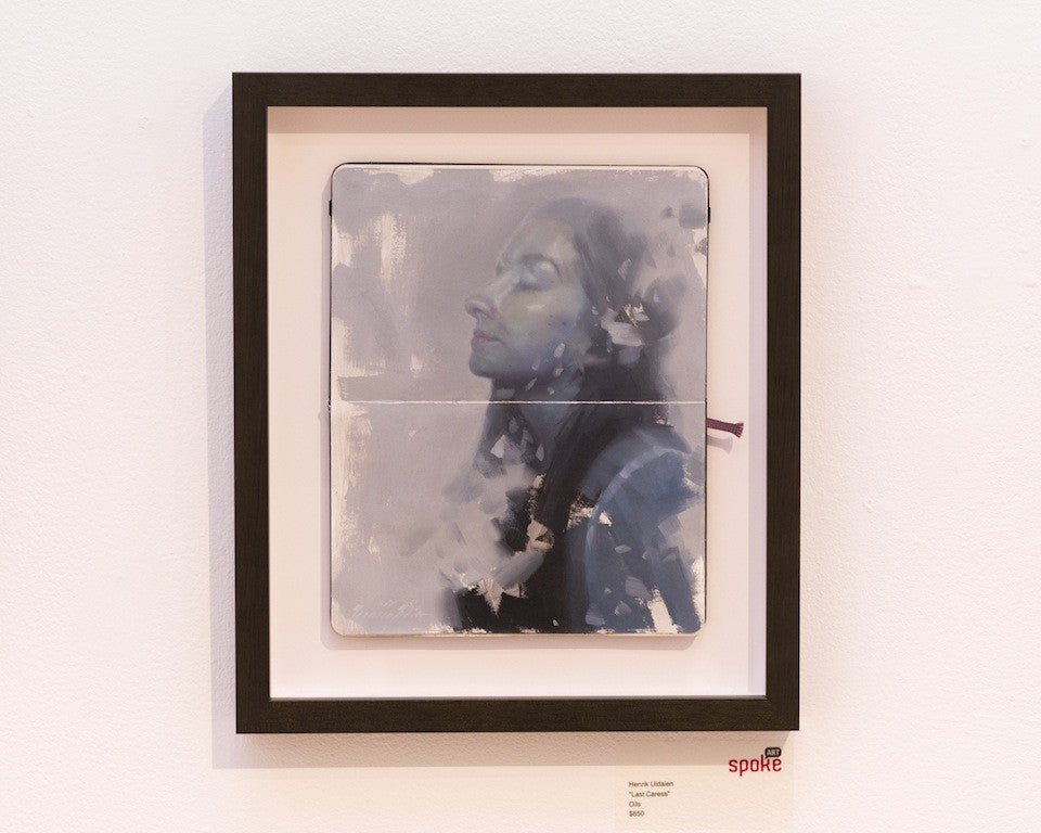 A framed artwork titled Last Caress by Henrik Uldalen features a portrait of a person with closed eyes, painted in the artists signature soft, abstract style using muted colors. Its an oil on moleskine piece accompanied by a small label beneath the frame.