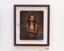 A framed oil painting by Crystal Chan shows a young woman with dark hair on a bed in a white tank top. The dim light highlights her thoughtful expression, beautifully set against a white wall. Available for purchase.