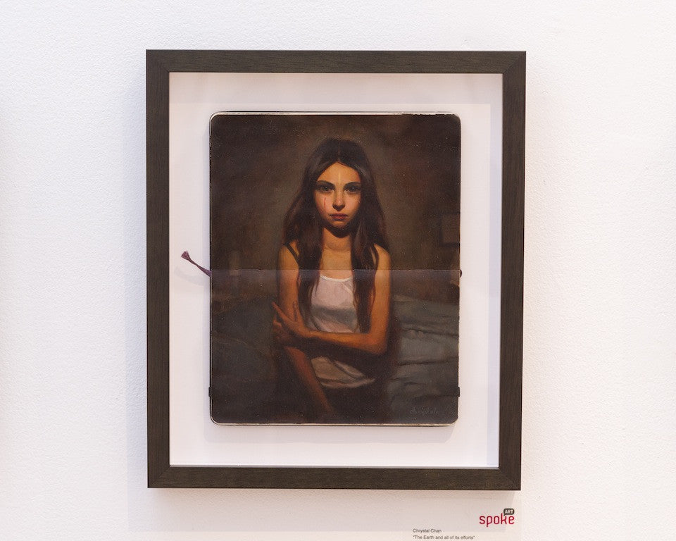 A framed oil painting by Crystal Chan shows a young woman with dark hair on a bed in a white tank top. The dim light highlights her thoughtful expression, beautifully set against a white wall. Available for purchase.