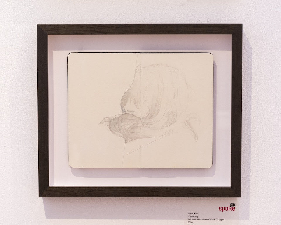 A Steve Kim framed graphite pencil drawing displays a persons partially visible head from behind, featuring long, flowing hair. Its mounted on a white wall and set within an open notebook.