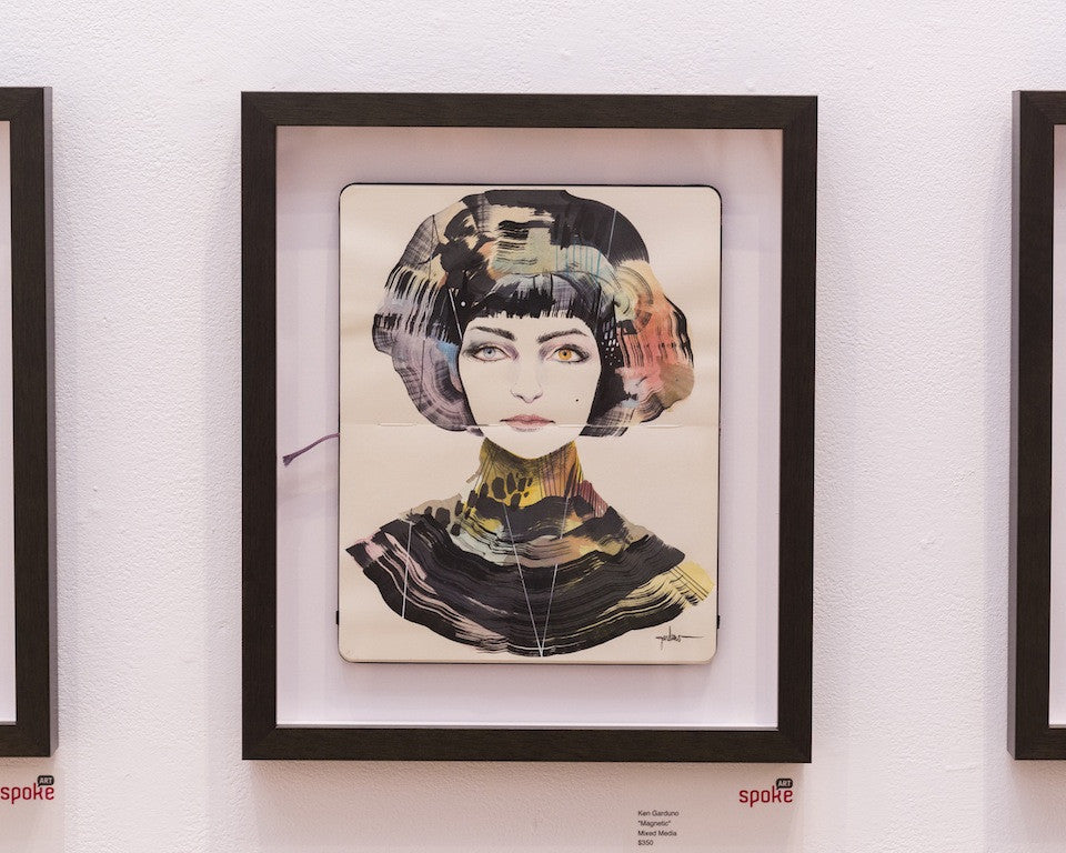 This magnetic mixed media artwork by Ken Garduno showcases a stylized portrait of a woman with short, multicolored hair and exaggerated eyes, featuring abstract brushstrokes in muted colors. Its displayed on a pristine white wall.