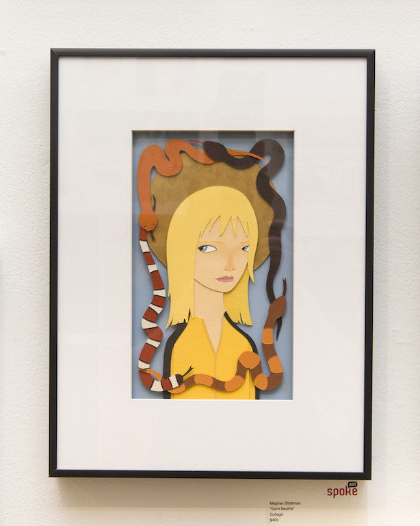 Meghan Stratmans framed artwork shows an animated character with blonde hair in a yellow outfit, surrounded by two snakes—one red with white stripes and one brown—against a textured collage-like background, displayed on a white wall.