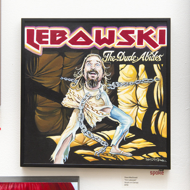 Dave MacDowells Iron Lebowski illustration depicts a bearded man with long hair, wrapped in chains and a straitjacket. The piece features the words LEBOWSKI and The Dude Abides, set against a dramatic black and yellow acrylic canvas background, elegantly framed on a white wall.