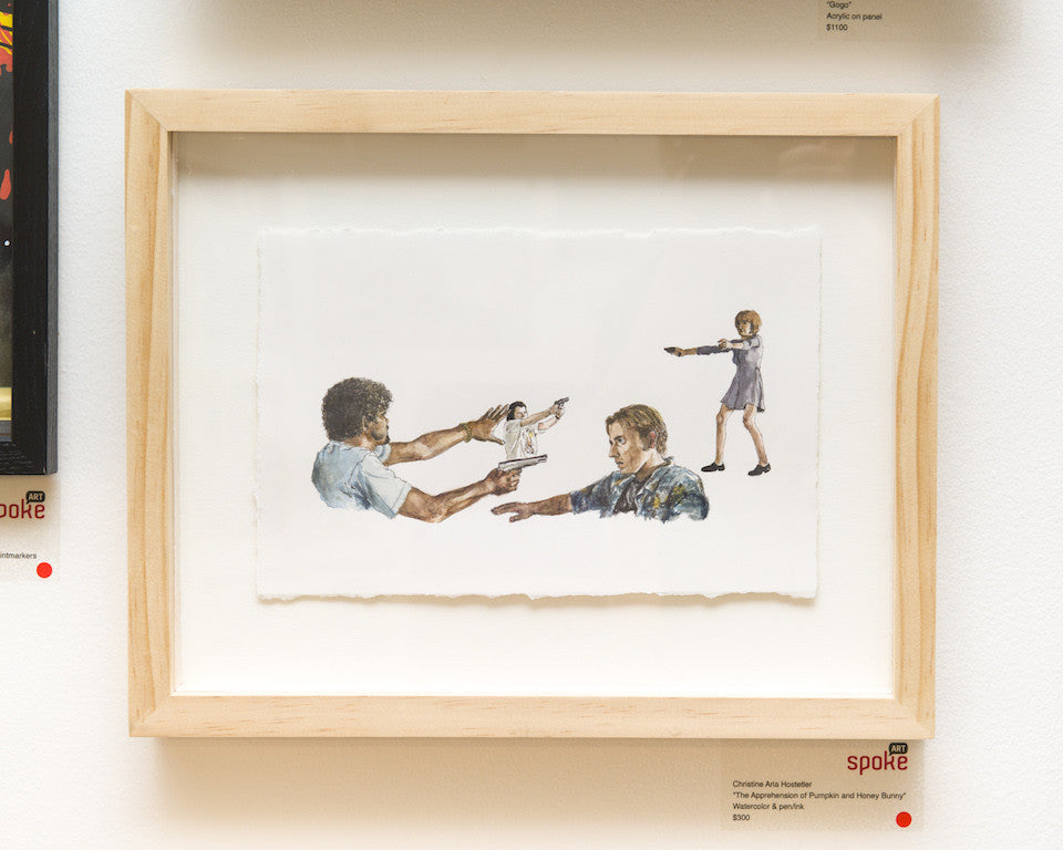 A Christine Hostetler framed watercolor shows a man pointing a gun at another, who surrenders his wallet, while a child in the background targets the first man. The artwork, reminiscent of Quentin vs Coen style, is displayed on a gallery wall in its wooden frame.