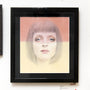 Bec Winnels framed artwork Uma features a surreal portrait of a short-haired woman, drawn with pencil and pastel on paper. The background is peach above and pale yellow below, while her eyes exude a soft, dreamy expression.