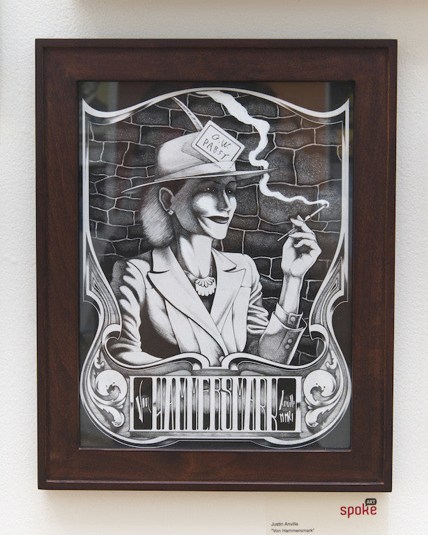 A striking ink on paper artwork by Justin Anville features a person in a hat and suit with a cigarette. NOIR is inscribed on the hatband against a brick backdrop. This framed piece is displayed on the wall with a small label beneath it.