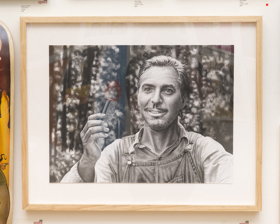 A black and white framed drawing by Joel Phillips shows a smiling person in overalls with a comb, set against blurred outdoor trees, crafted expertly with charcoal and graphite on paper.