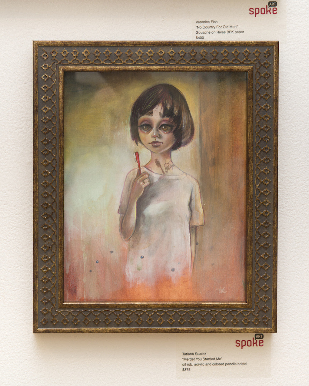 A framed Tatiana Suarez painting features a young person with short dark hair and big eyes, holding a red stick against a warm-hued background. Titled Merde! You Startled Me, its showcased on a white wall with price tags below.