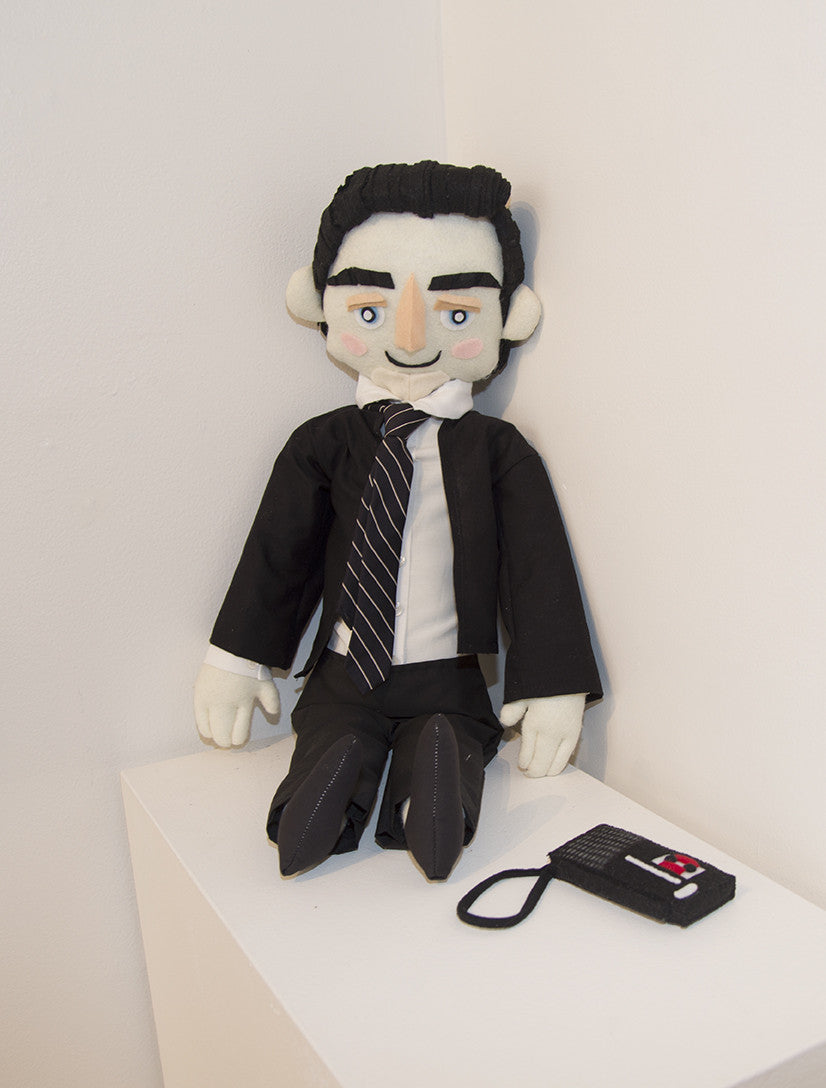 A plush FBI Special Agent doll with black hair, a suit, tie, and shoes leans on a white wall. Displaying Tessa Morrisons exquisite fiber art with a small smile, it sits near a small black and red device with a strap.