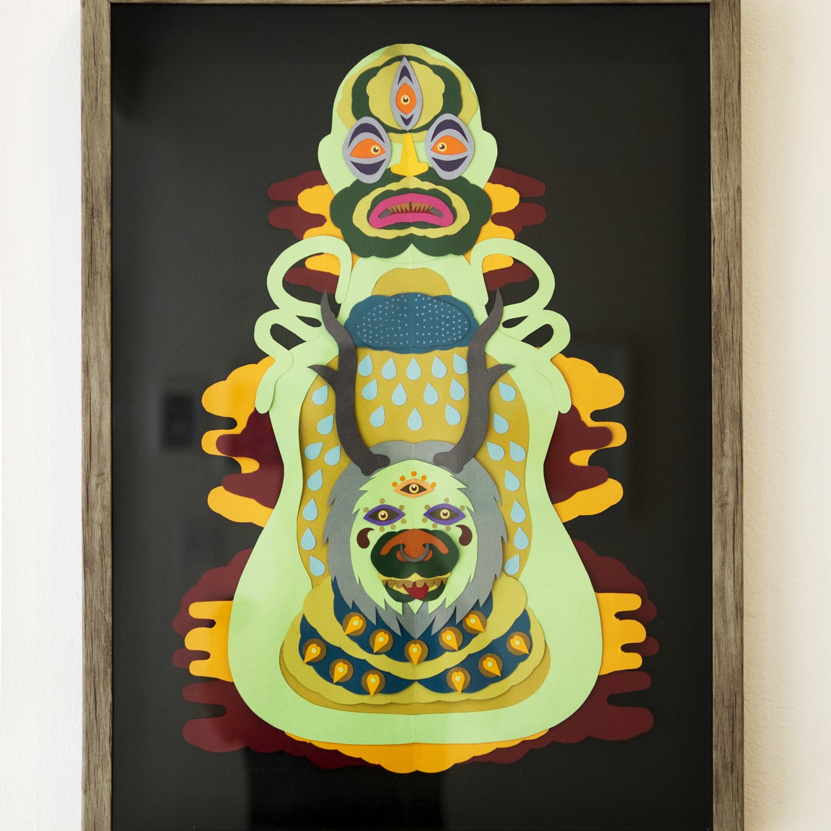 Daniel Jaffes framed abstract artwork features a complex, symmetrical design with vibrant colors. The layered paper art depicts face-like shapes with multiple eyes and mouths in green, yellow, blue, and red hues on a dark background.