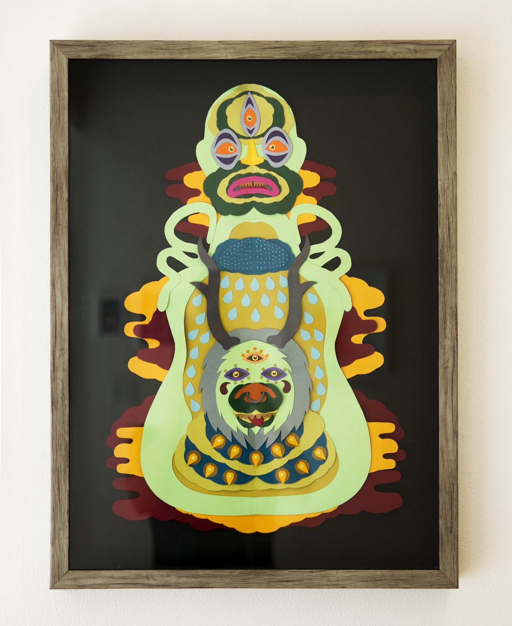 Daniel Jaffes framed abstract artwork features a complex, symmetrical design with vibrant colors. The layered paper art depicts face-like shapes with multiple eyes and mouths in green, yellow, blue, and red hues on a dark background.