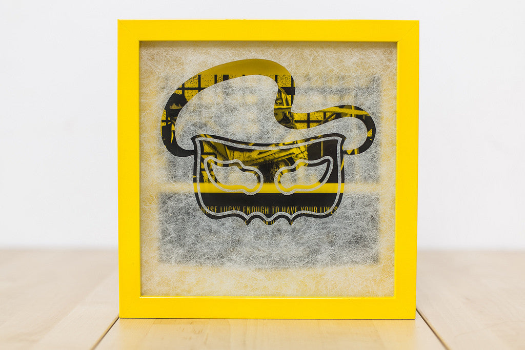 A stylized black eye mask with swirling hat details, inspired by Derek Ballards style, is set against a textured white background. The ink artwork on transparent film is framed in bright yellow and elegantly rests on a light wooden surface.