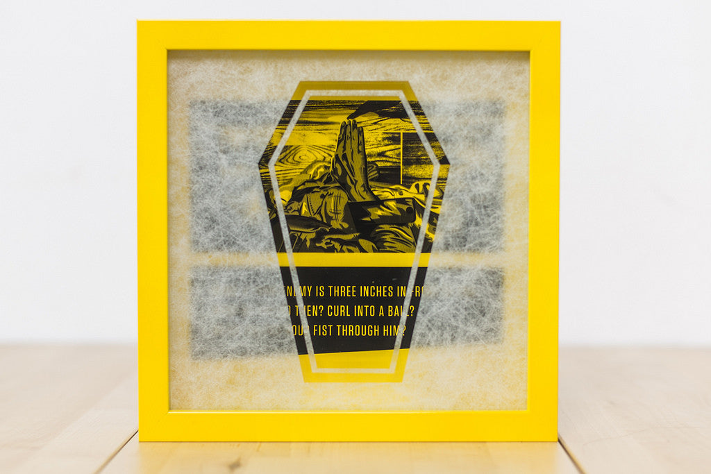 Derek Ballards framed art features a stylized hand amid a landscape with a yellow border and partially obscured text appearing like its behind a rice paper stencil, all on a light wooden surface.