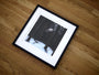 A framed fine art giclee print rests on a wooden floor, depicting a snowy forest with tall, dark trees. A hand emerges among the trees, evoking Henrik Uldalens mystique—a timeless piece expertly preserved through archival framing by the Henrik Uldalen brand.