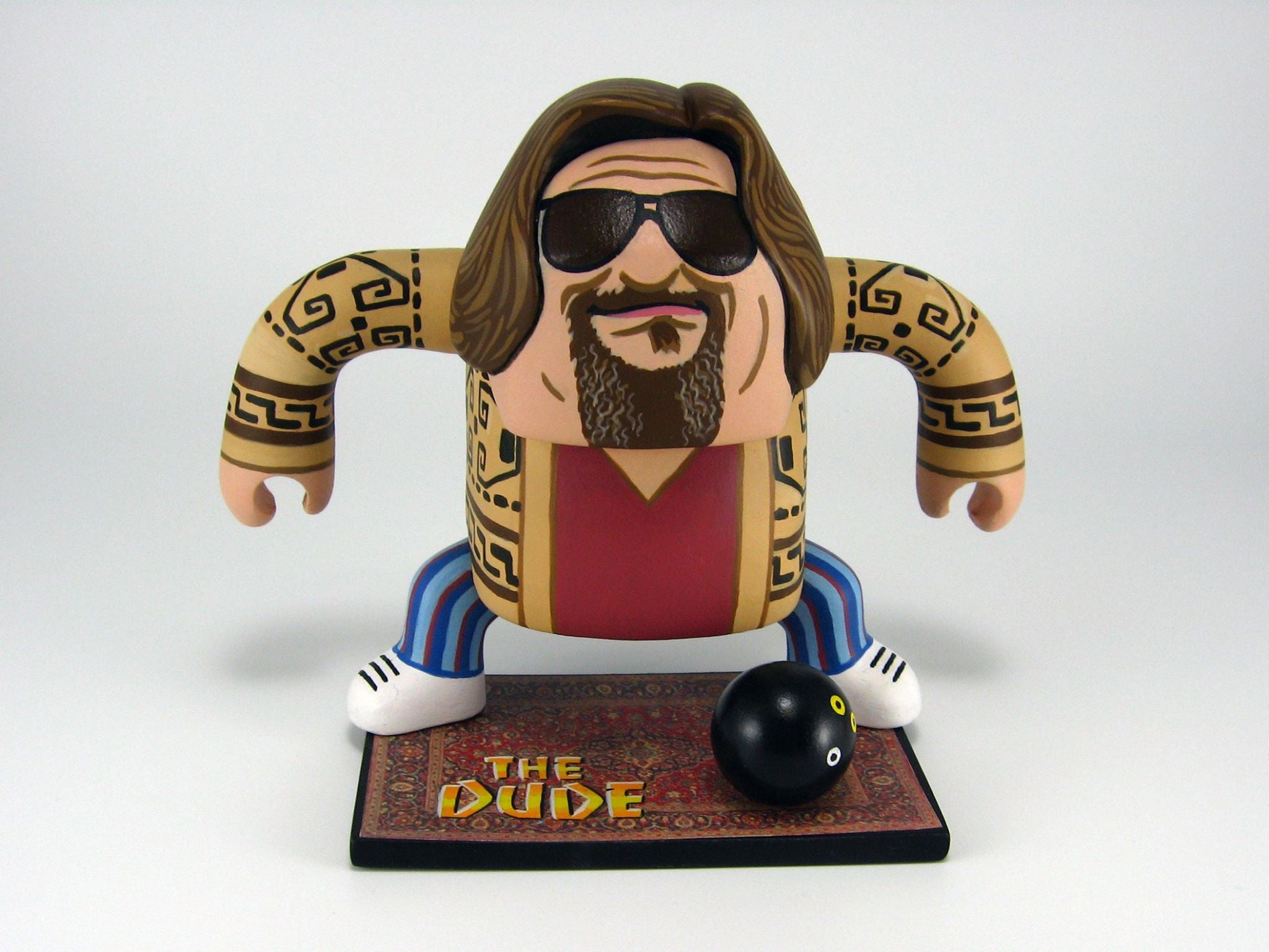 A cartoon figure of a long-haired, bearded man wearing sunglasses, a patterned sweater, and striped pants stands on a The Dude vinyl mat with a bowling ball in front. Brand: Geoff Trapp.