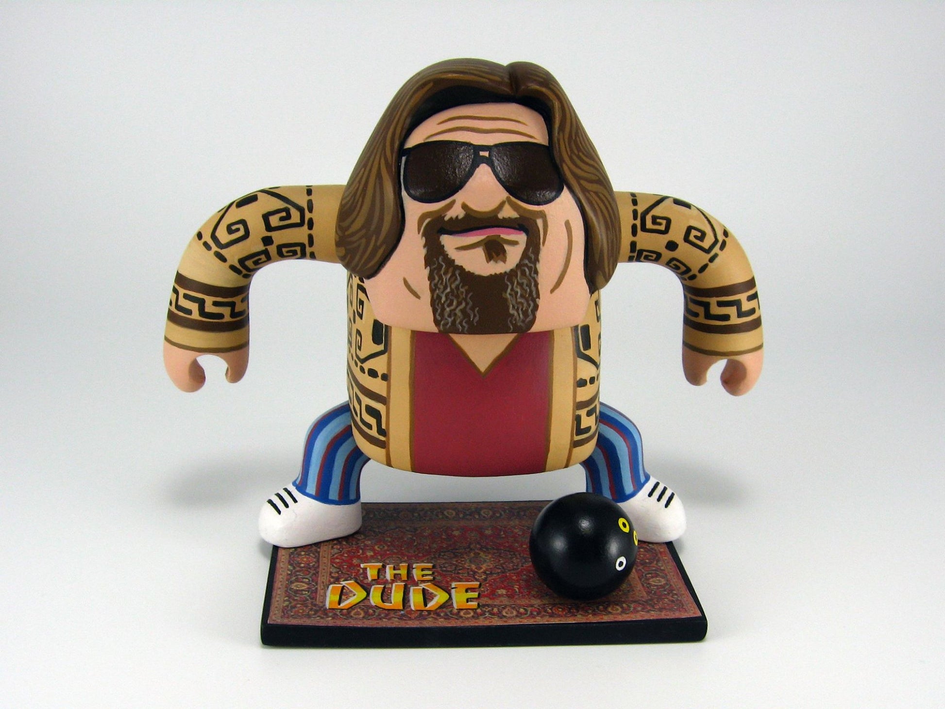A cartoon figure of a long-haired, bearded man wearing sunglasses, a patterned sweater, and striped pants stands on a The Dude vinyl mat with a bowling ball in front. Brand: Geoff Trapp.