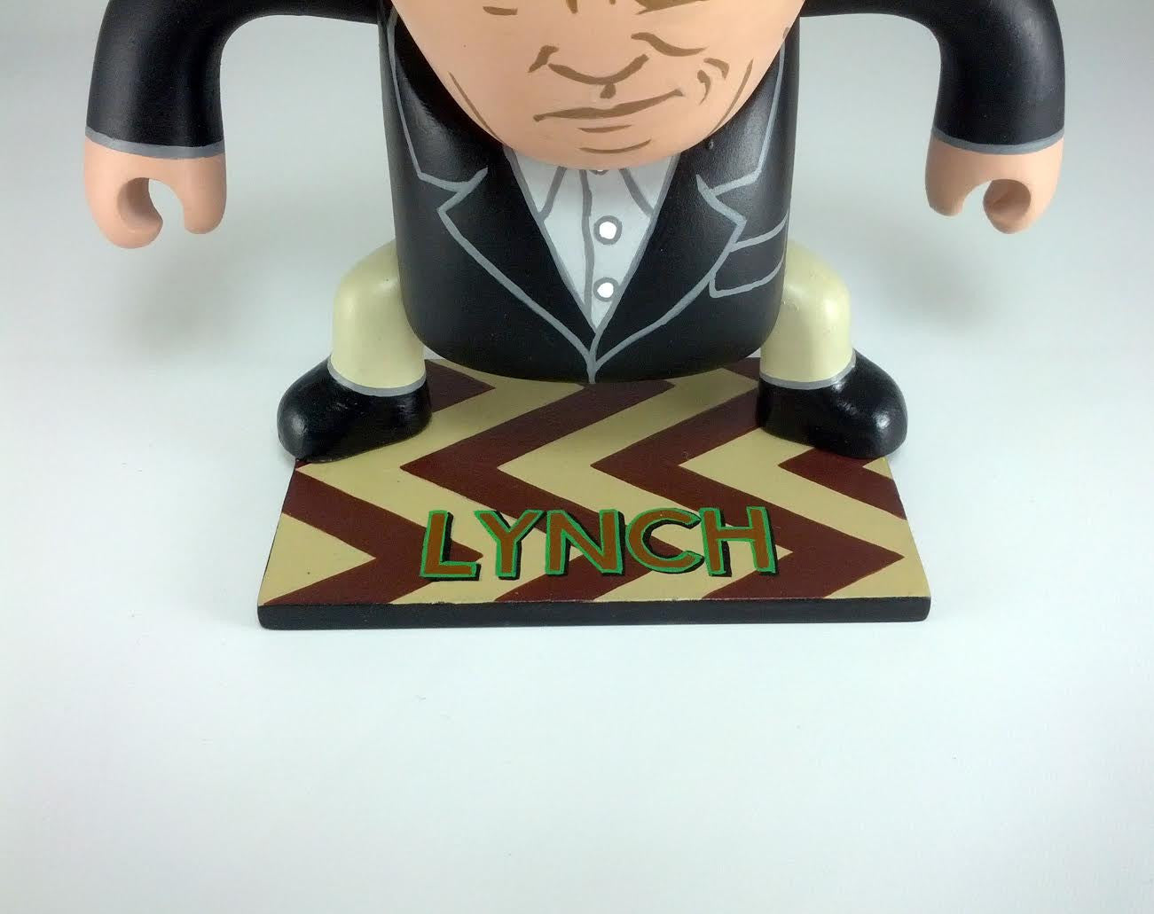 A Vinyl Sculpy figurine by Geoff Trapp stands on a patterned base with LYNCH in green letters. Showcased at the Spoke Art Gallery, it features a suit and exaggerated features against a plain white background.