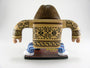 A stylized figure with long hair stands on a vinyl-like platform, wearing a tan shirt with tribal patterns and blue-striped pants. Arms are outstretched, capturing Geoff Trapps mixed media style.