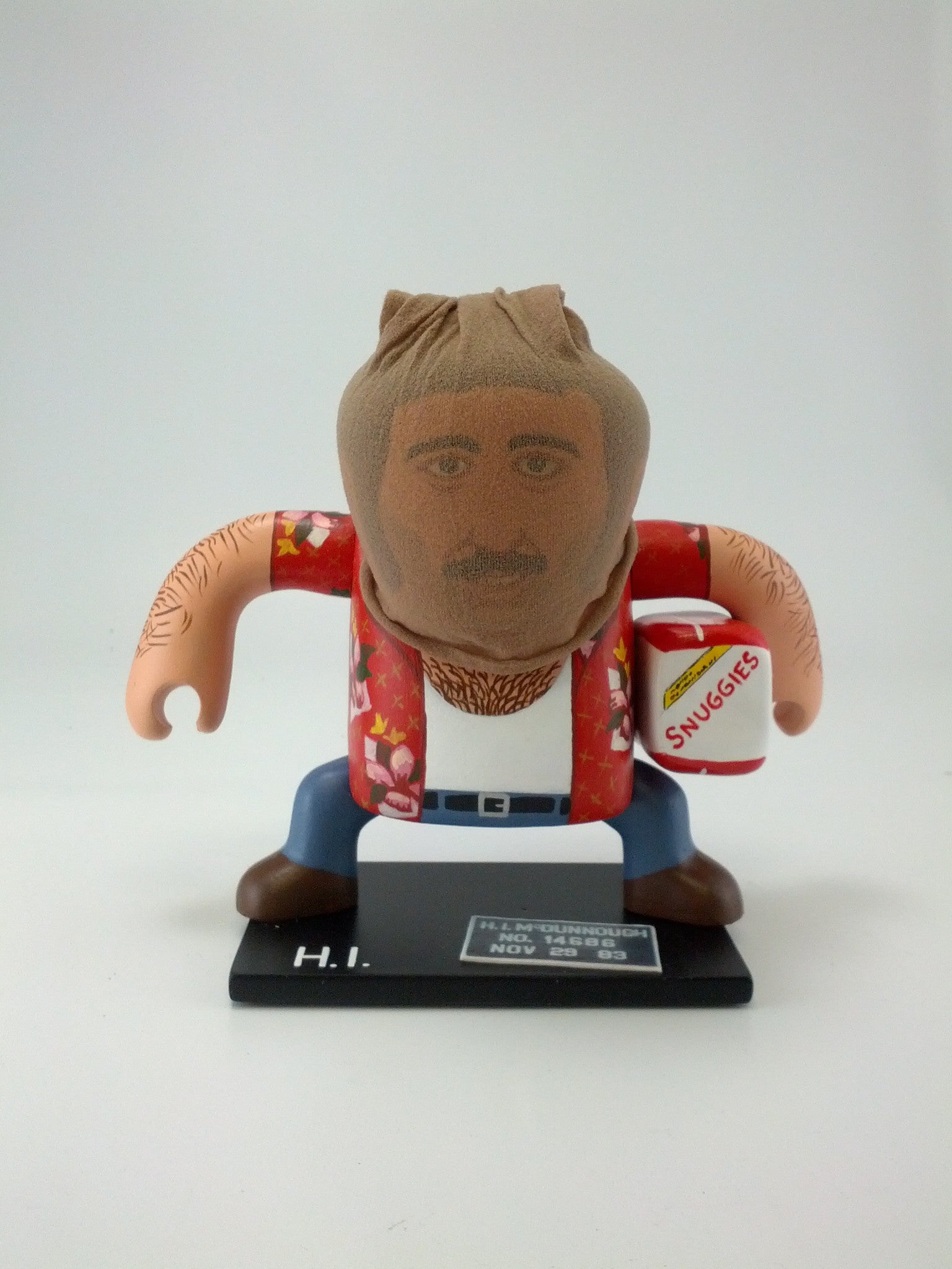 A Geoff Trapp toy figure, exuding playful charm, wears a floral shirt and blue pants on an H.I. base. Its head is wrapped in nylon stocking while holding a Huggies Snuggies package.