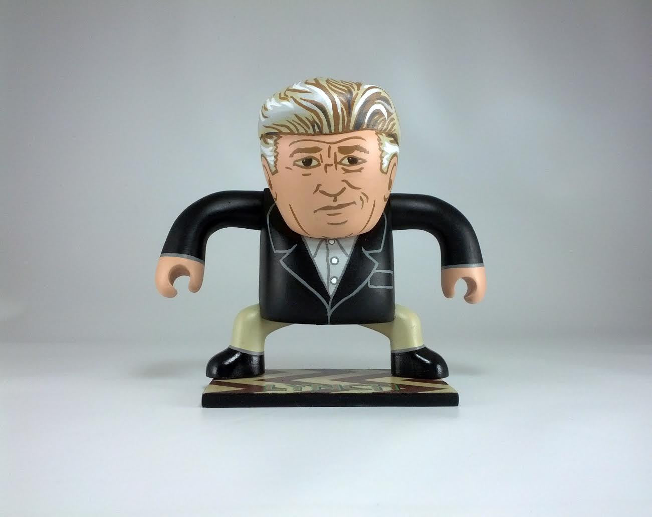A vinyl Geoff Trapp caricature toy features exaggerated facial details, a large head, stylized hair, and a suit. It resembles a cartoonish older figure in formal attire with its base stand.