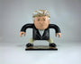 A vinyl Geoff Trapp caricature toy features exaggerated facial details, a large head, stylized hair, and a suit. It resembles a cartoonish older figure in formal attire with its base stand.