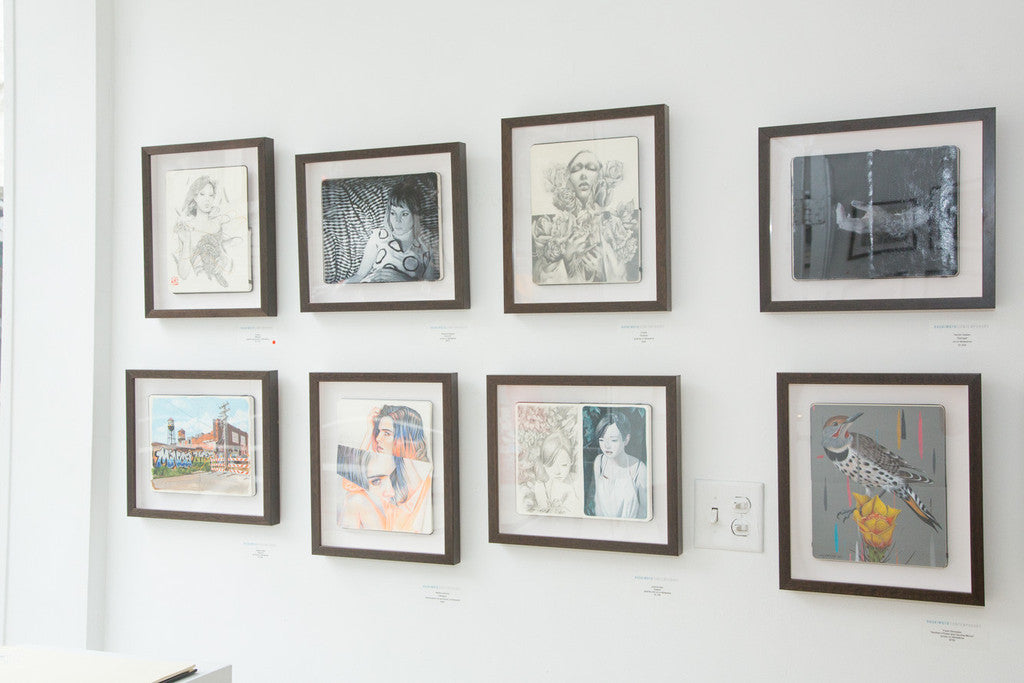 The gallery wall showcases eight custom framed artworks by Jessica Hess, featuring sketches, paintings, and photos. Highlights include portraits, abstracts, a colorful bird, and a striking gouache on Moleskine. Displayed in a modern white room with a small control panel.