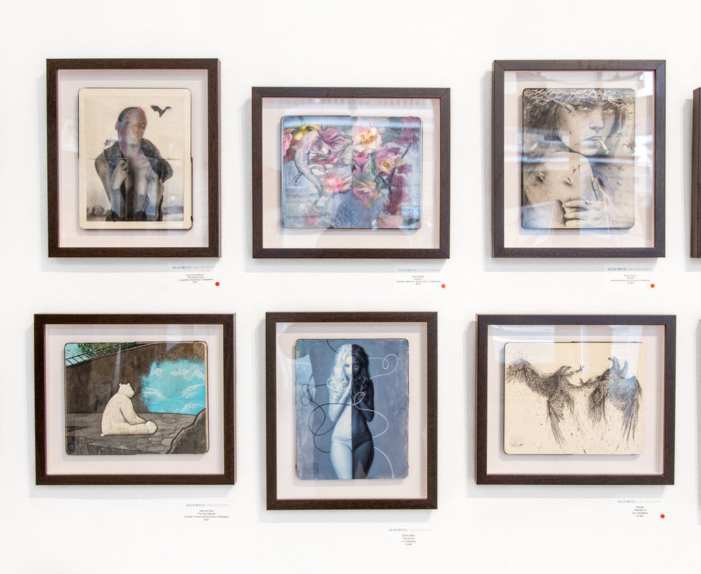 In a gallery, a wall display features six framed artworks captivating viewers with Brian M. Viveros-inspired portraits and abstract designs, including a stunning charcoal piece. Each artwork has a small descriptive card beneath to share its unique story.