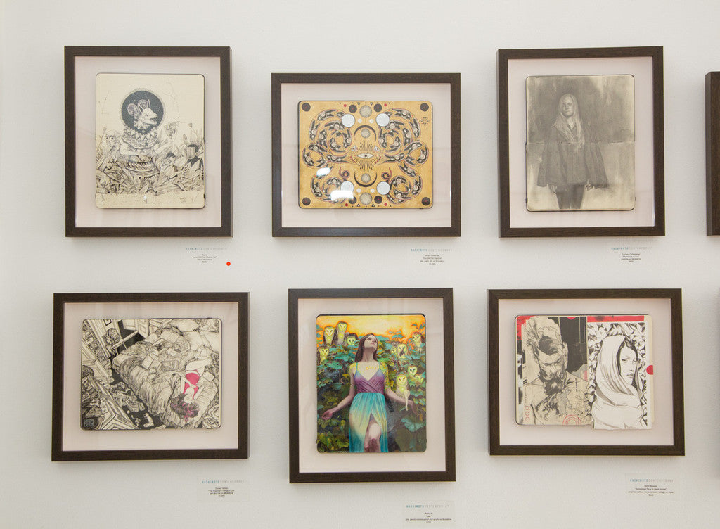 A gallery wall with six framed artworks showcases diverse styles: a detailed Moleskine sketch, an abstract piece echoing Saners style, and a vibrant Cosmic Girl portrait. Frames are evenly spaced on an immaculate white wall.