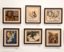 A gallery wall displays six framed artworks, including Reinier Gamboa portraits, abstract designs, and nature scenes. Among them is Ram Sees the Third, crafted in graphite on Moleskine. The pieces are arranged in two elegant rows of three, highlighting diverse styles and colors.
