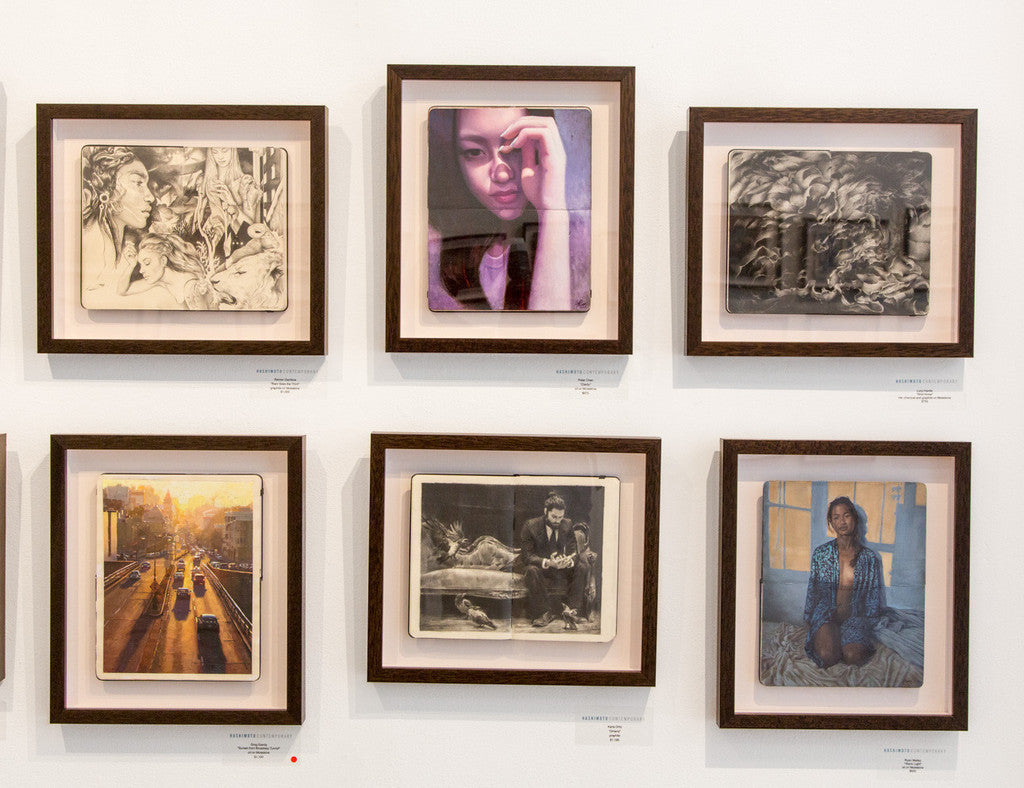 A wall features six framed photographs in two rows, showcasing Peter Chans meticulous style with portraits and realistic scenes reminiscent of oil on Moleskine artistry, including a sitting woman and a cityscape, all framed in dark wood.