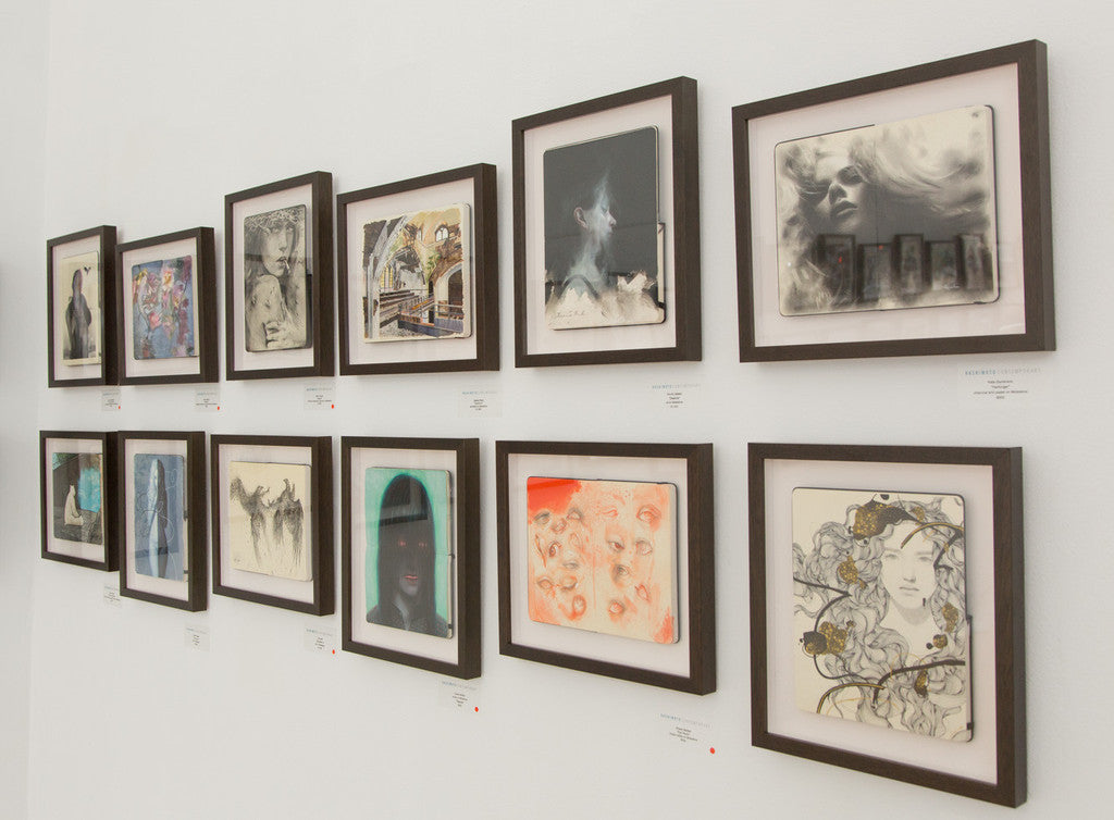 A gallery wall showcases multiple framed artworks by Kate Zambrano, featuring portraits and abstract imagery. Dark frames contrast against a light wall. Each drawing, crafted with charcoal and pastel, has an informative label beneath it.