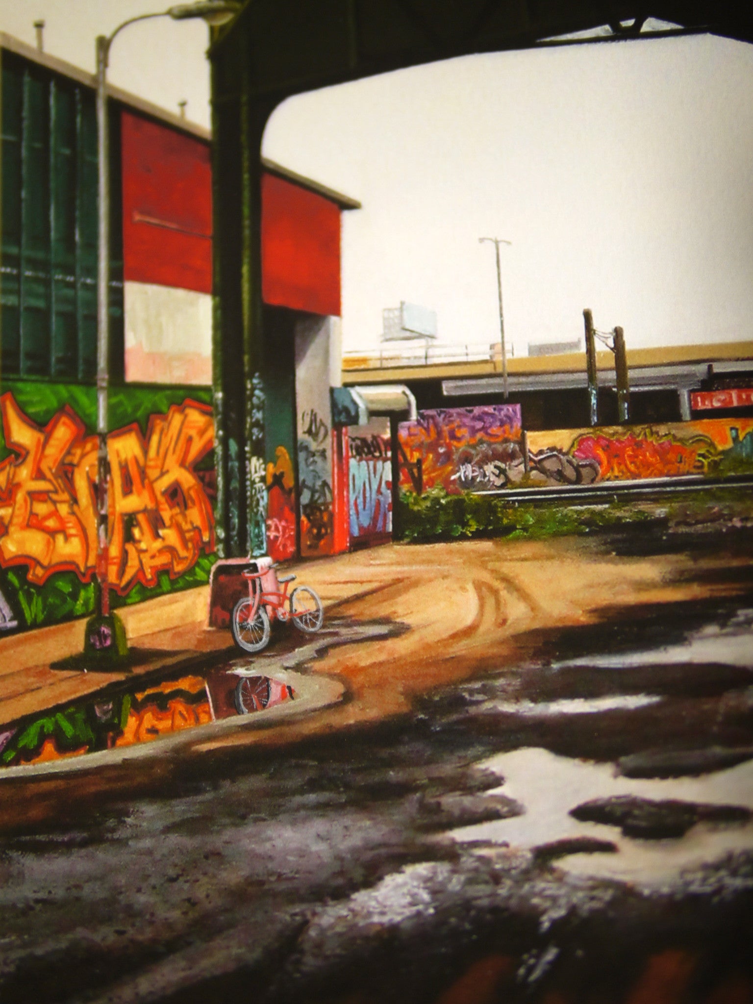 Amid a vibrant street scene with colorful graffiti, a solitary red bicycle rests by a reflective puddle on worn pavement. The urban setting, enhanced by industrial structures and an overpass, feels like a Jessica Hess hand-painted piece brought to life with archival Epson Ultrachrome HDR inks.