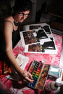 A person with short dark hair and glasses paints at a table covered in a red patterned cloth, surrounded by cityscape prints. They use archival Epson Ultrachrome HDR inks from the Jessica Hess watercolor set, enhancing the artistic and focused atmosphere.