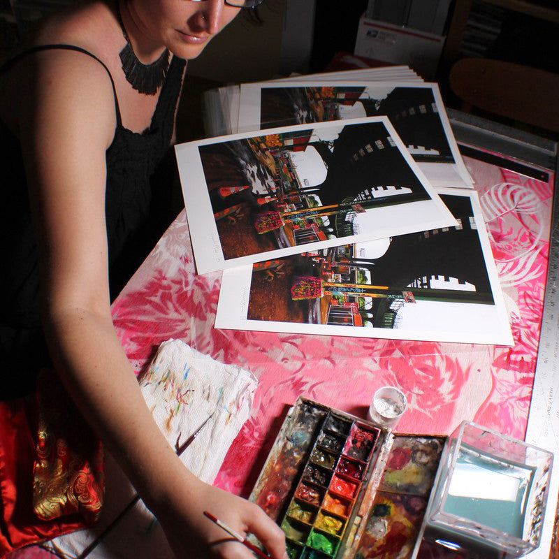 A person with short dark hair and glasses paints at a table covered in a red patterned cloth, surrounded by cityscape prints. They use archival Epson Ultrachrome HDR inks from the Jessica Hess watercolor set, enhancing the artistic and focused atmosphere.
